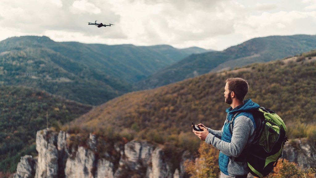 A person standing on a hilltop operates a drone against a backdrop of mountains and cloudy skies.