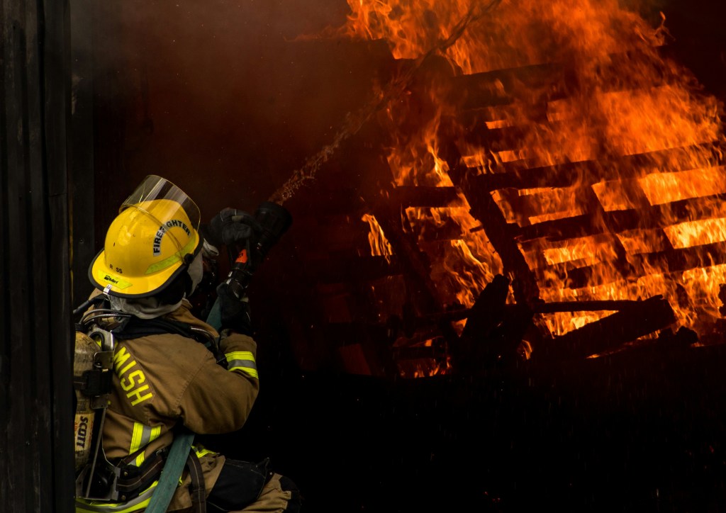 A firefighter in full protective gear directs a stream of water at a large blaze consuming a stack of wooden pallets.
