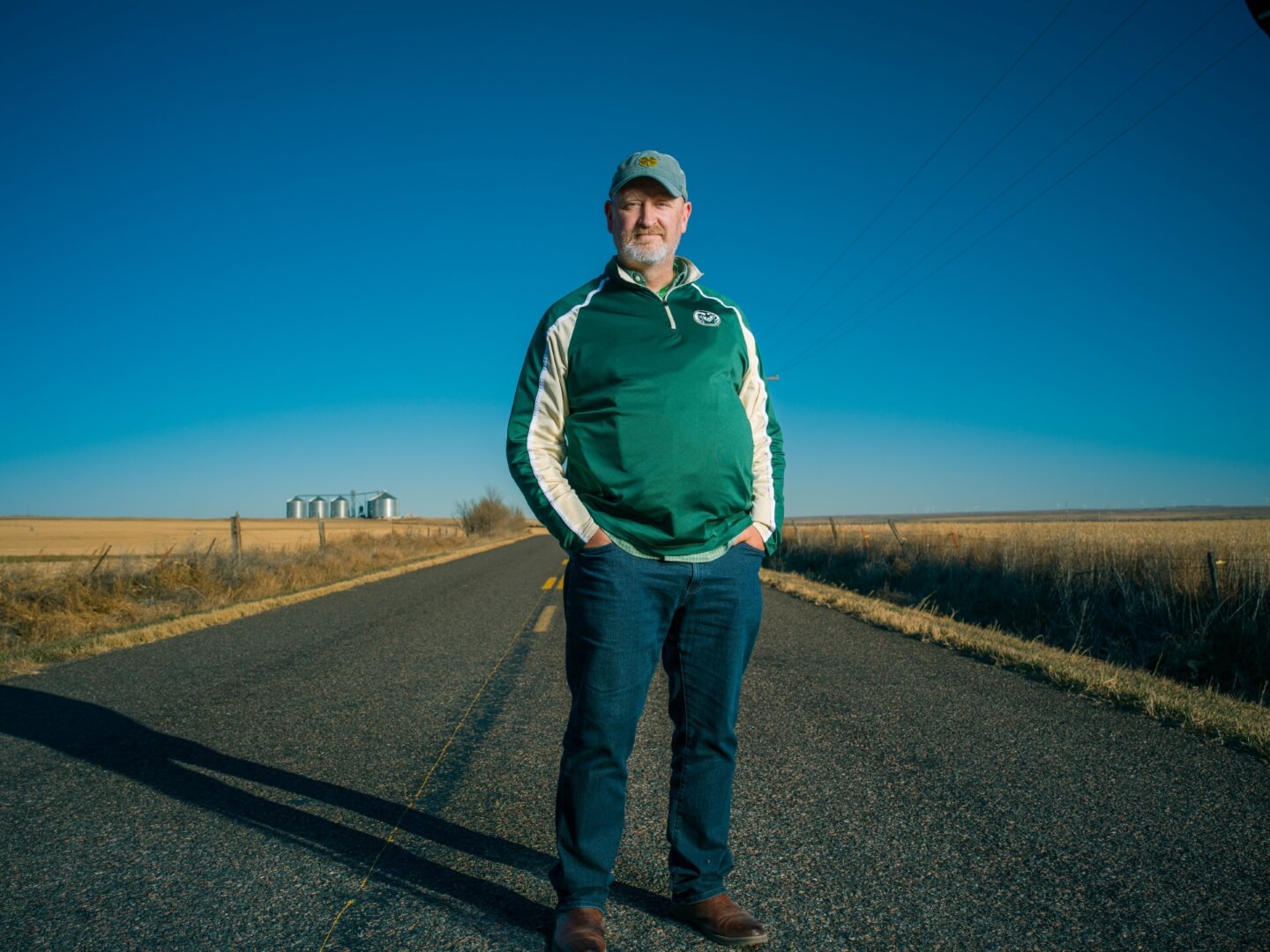 A person standing on a rural road with hands in pockets, wearing a green jacket and cap. The background shows open fields, a clear blue sky, and grain silos in the distance.