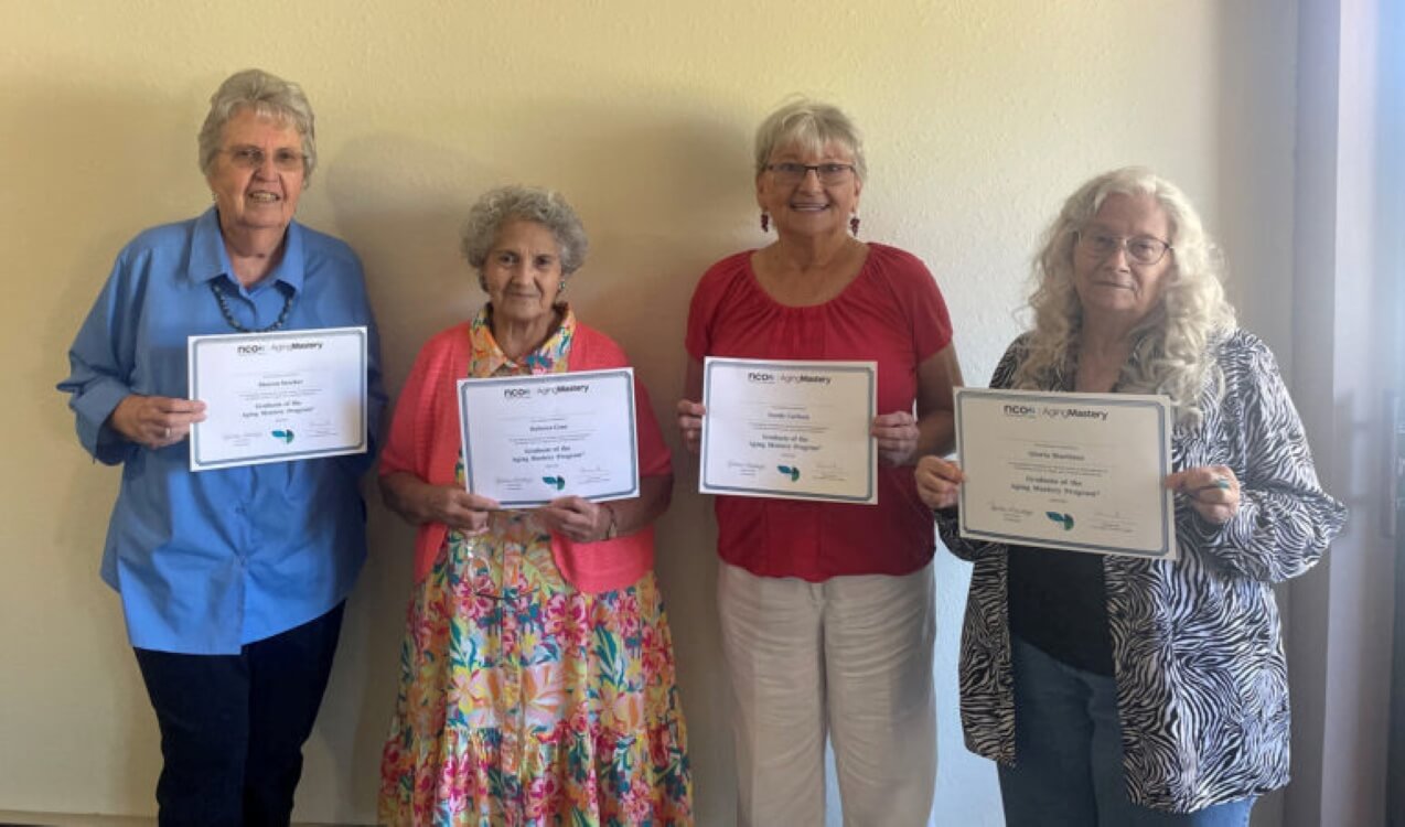 Four people standing indoors holding certificates in front of a light-colored wall. The certificates have text and logos visible, indicating an achievement or completion of a program.