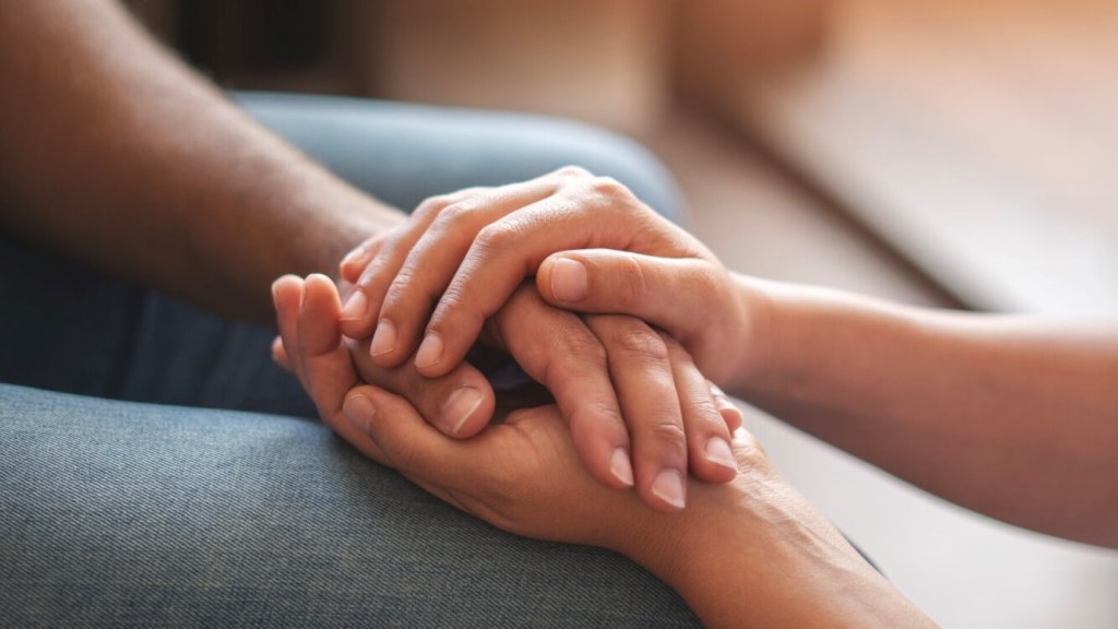 Close-up of two hands gently clasping each other, suggesting comfort or support. The background is softly blurred.