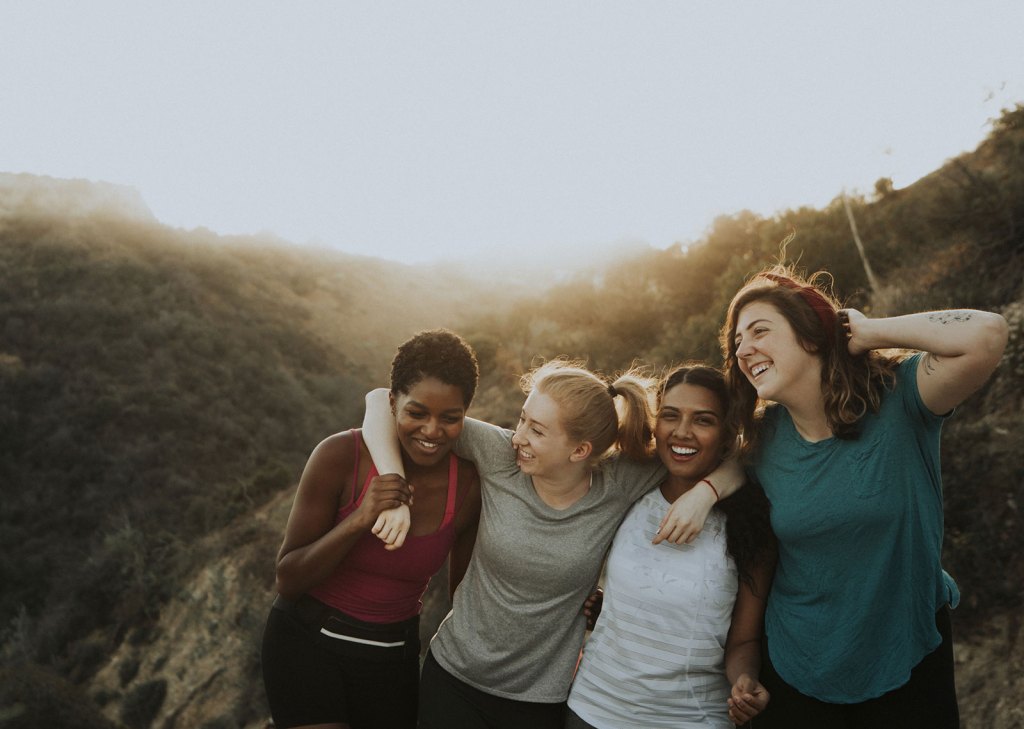 Four women smiling and embracing each other during a hike, with hills and the sun in the background.