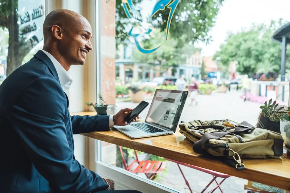 A man sitting at a café window with a laptop and phone, smiling while working, with a view of an outdoor plaza in the background.