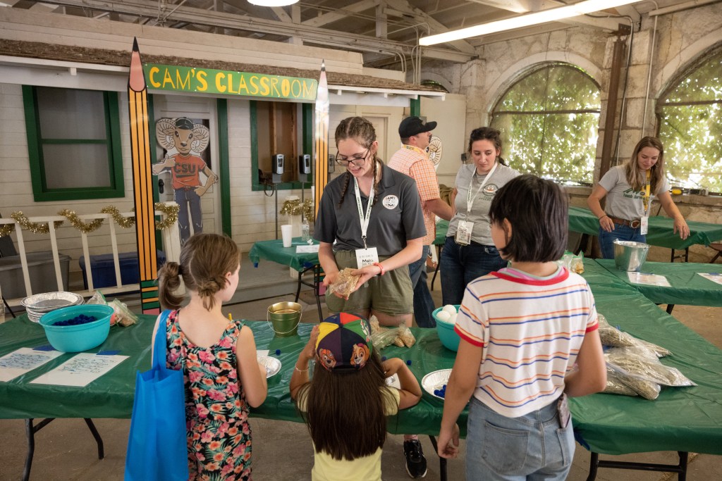 A group of children gathered around a table at an the Colorado State Fair, learning hands-on activities led by a guide in a room labeled "Cam's Classroom.