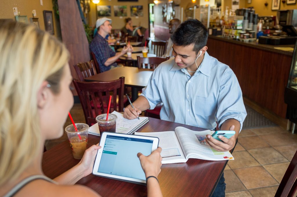 A man sits at a cafe table while listening to a lecture on his phone and taking notes.