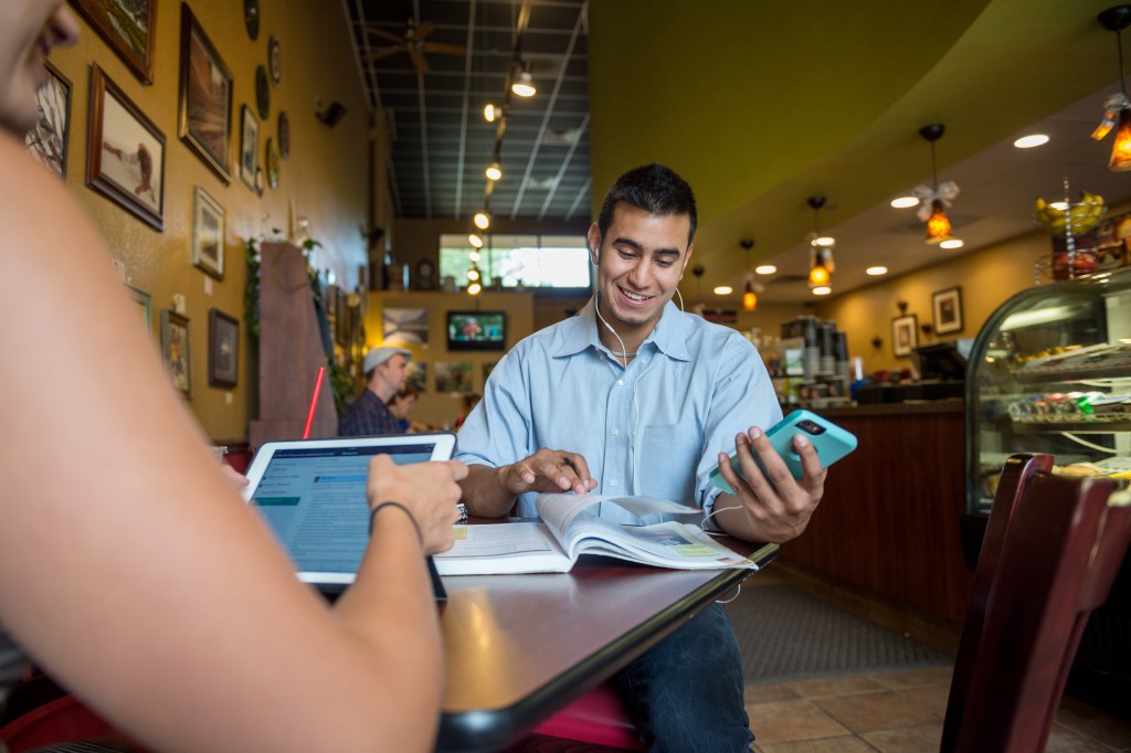 A man sits at a cafe table while listening to a lecture on his phone and taking notes.