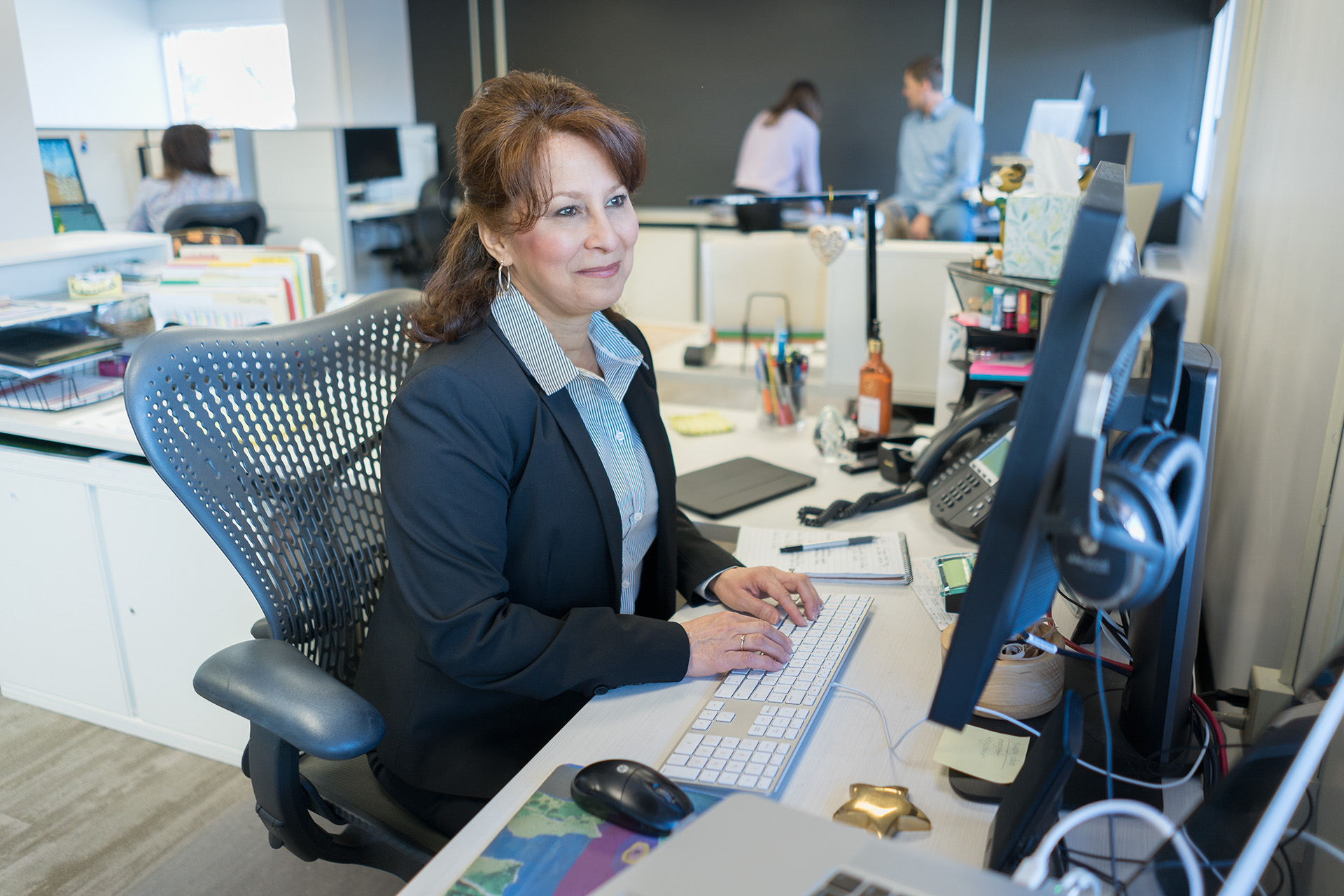 A person sitting in an office setting, looking thoughtfully as they listen, with colleagues working in the background.