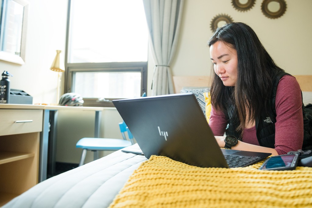 A person sitting on a bed in a dorm room, working on a laptop with a phone nearby.