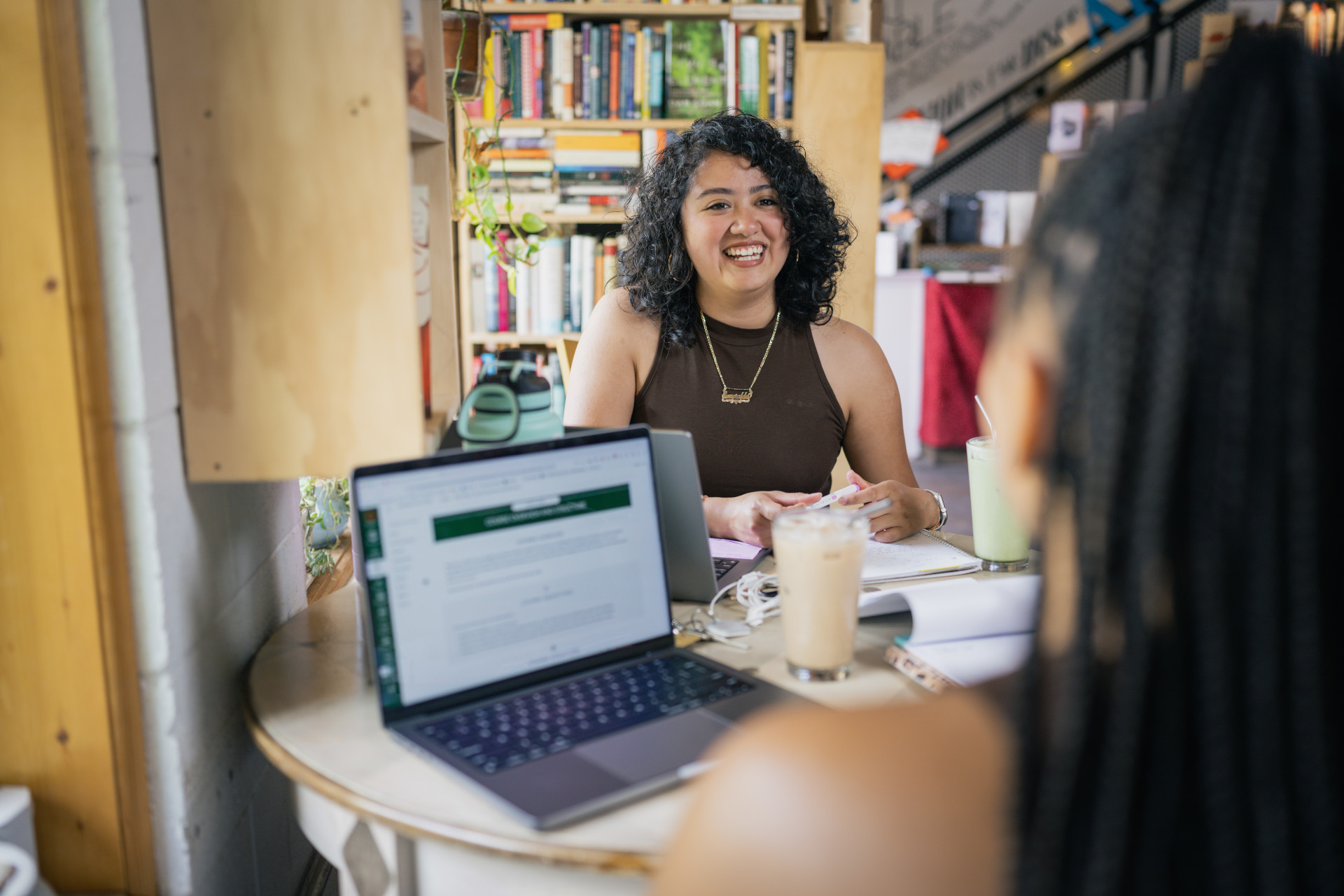 Two CSU students sitting at a table in a cozy café, smiling and working together, with a laptop and drink on the table.