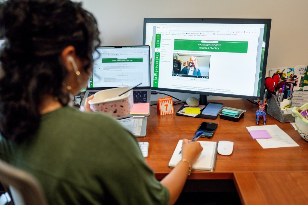 A person sitting at a desk, taking notes while participating in an online course on a computer with multiple screens. Various office supplies are scattered across the desk.