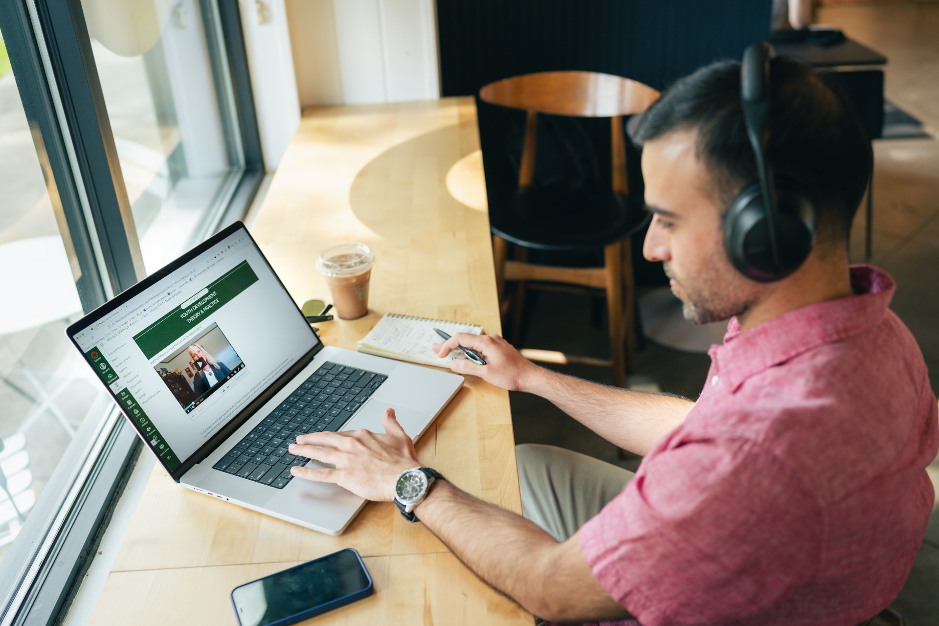 A person taking notes while watching an online course on a laptop, with headphones on and a coffee cup nearby.