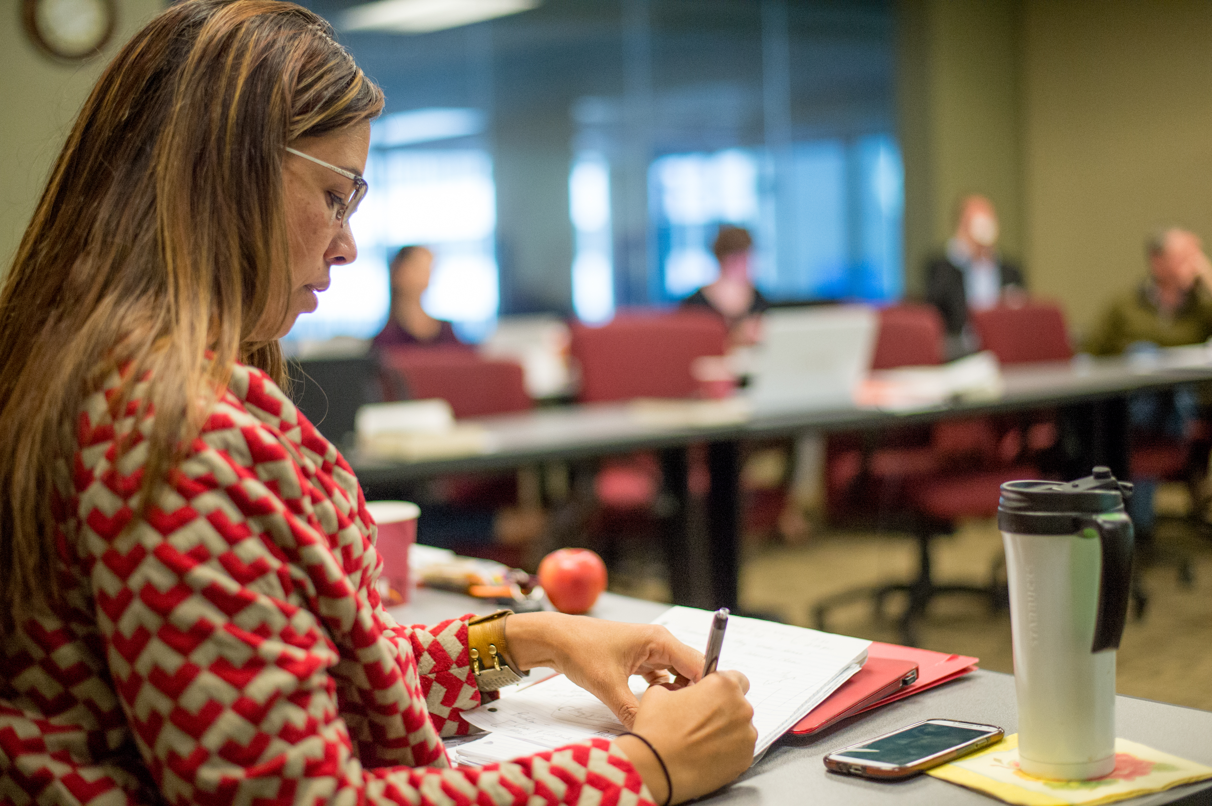 A woman sits in a classroom at a table with other students nearby.