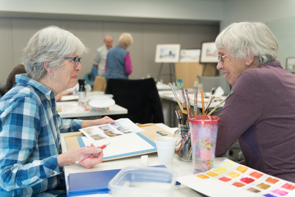 Two senior women smiling at each other during a watercolor painting class.