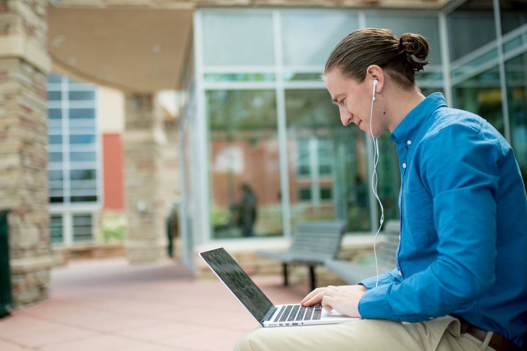 A person sitting outside on a bench, wearing earphones while working on a laptop.