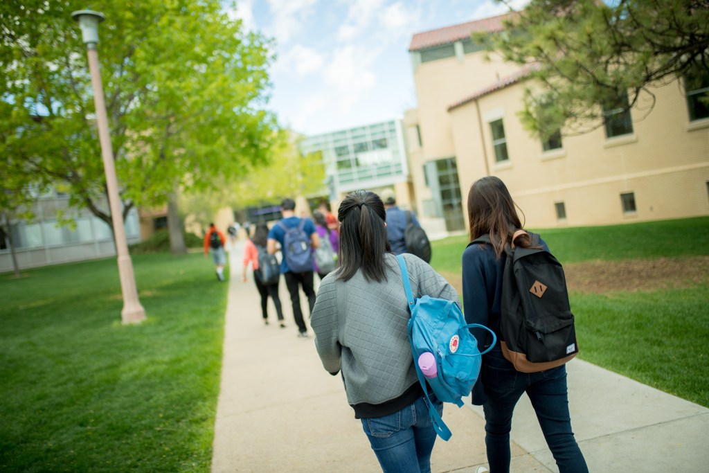 Two students with backpacks on walk down a sidewalk on campus.