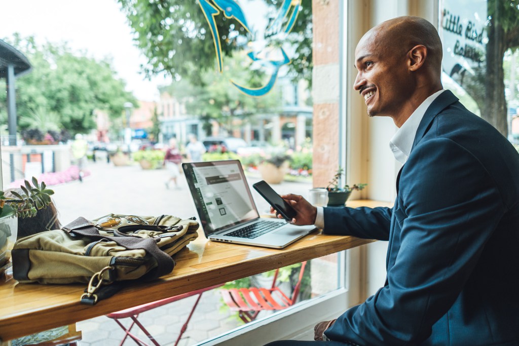 A man sitting at a café window with a laptop and phone, smiling while working, with a view of an outdoor plaza in the background.