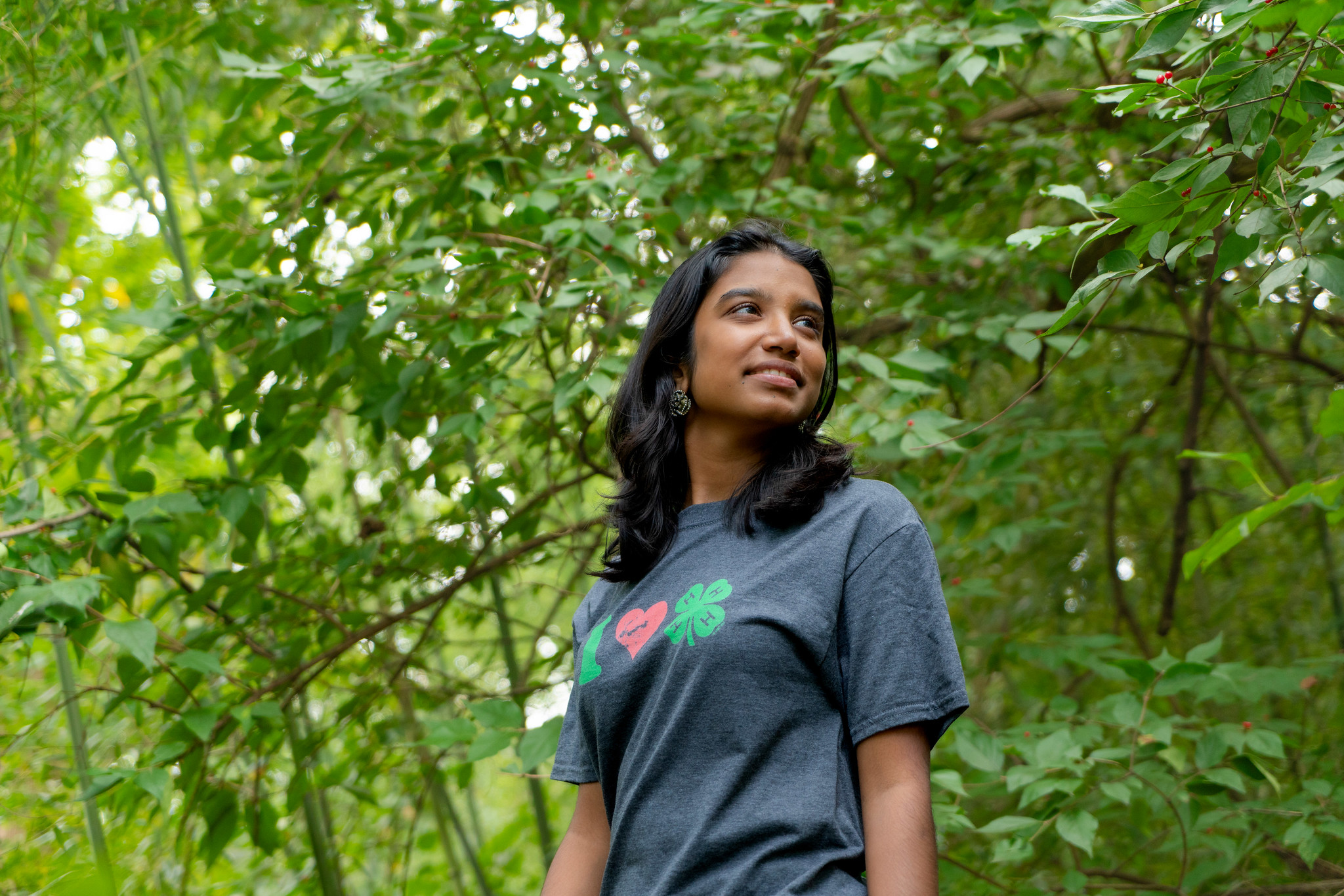 A person standing in a green, leafy environment, looking into the distance with a thoughtful expression.