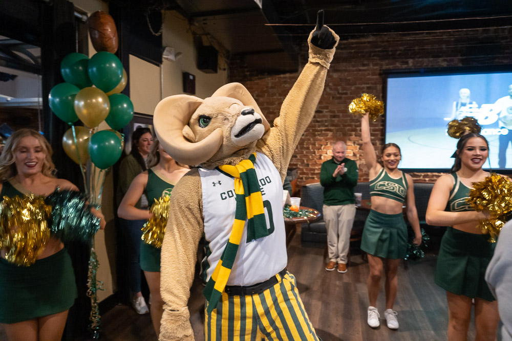 CAM the Ram, the Colorado State University mascot, raising a fist in celebration, surrounded by cheerleaders in a festive setting.