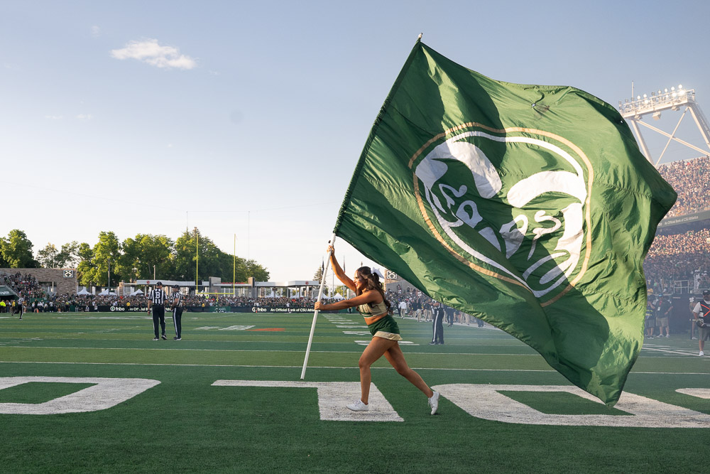 A CSU cheerleader running across CSU's football field holding a large CSU flag during a game.