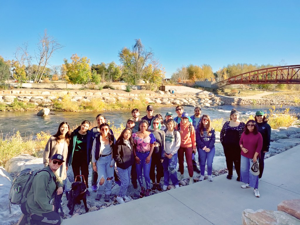 A group of students standing together near a river with a pedestrian bridge in the background.