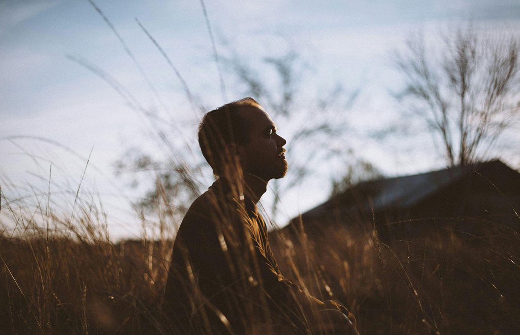 A man sitting outdoors in a field at sunset, meditating with his eyes closed.