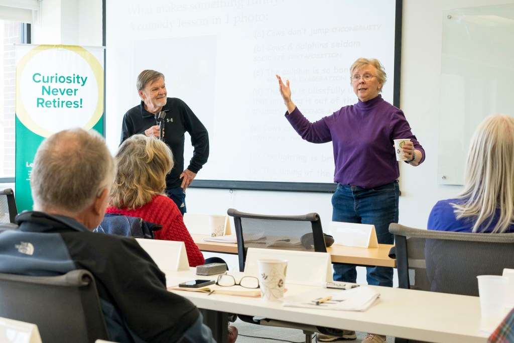 An older adult giving a presentation to a group of seniors seated in a classroom setting, with books and educational materials in the background.