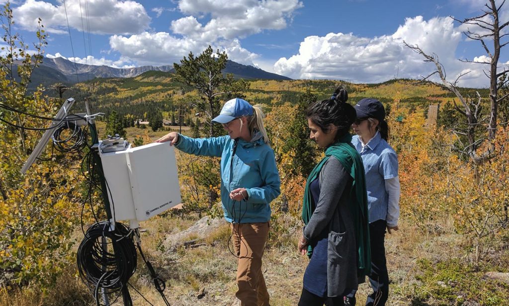 Three women conducting field research outdoors, with one woman adjusting scientific equipment in a mountainous, forested area under a bright, partly cloudy sky.