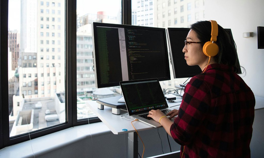 A person wearing headphones sits in front of two computer screens and a laptop.