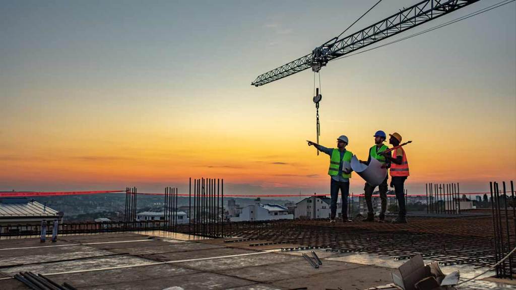 Construction workers in safety vests and helmets stand on a building site at sunrise, pointing and reviewing plans as a crane looms overhead.