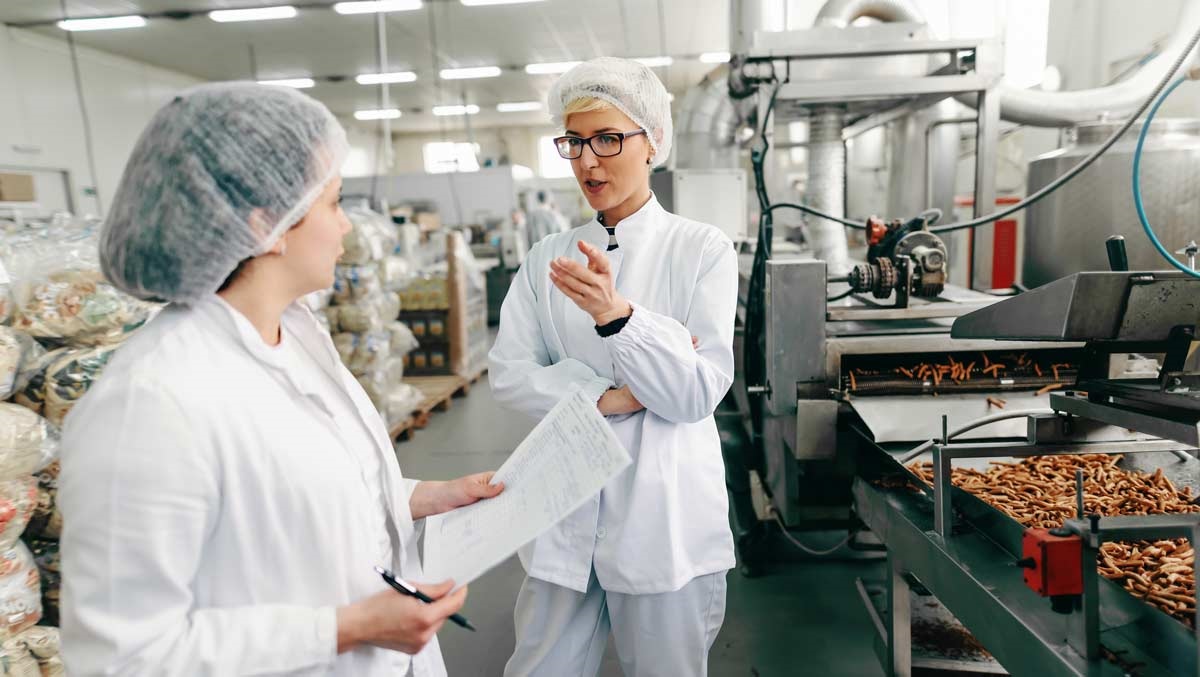 Two women wearing hairnets and lab coats have a discussion inside a food processing plant, surrounded by machinery and conveyor belts.