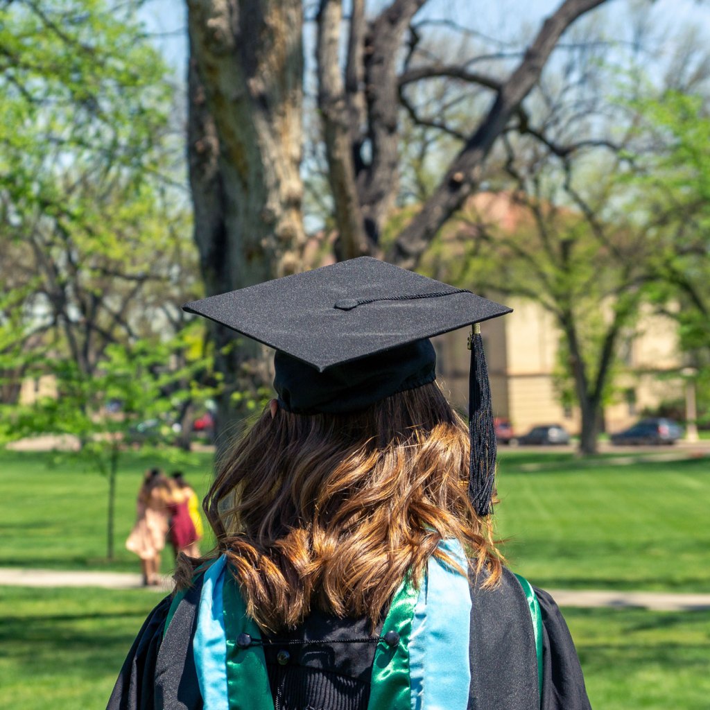 Graduate wearing a cap and gown with back facing the camera and trees in the background.