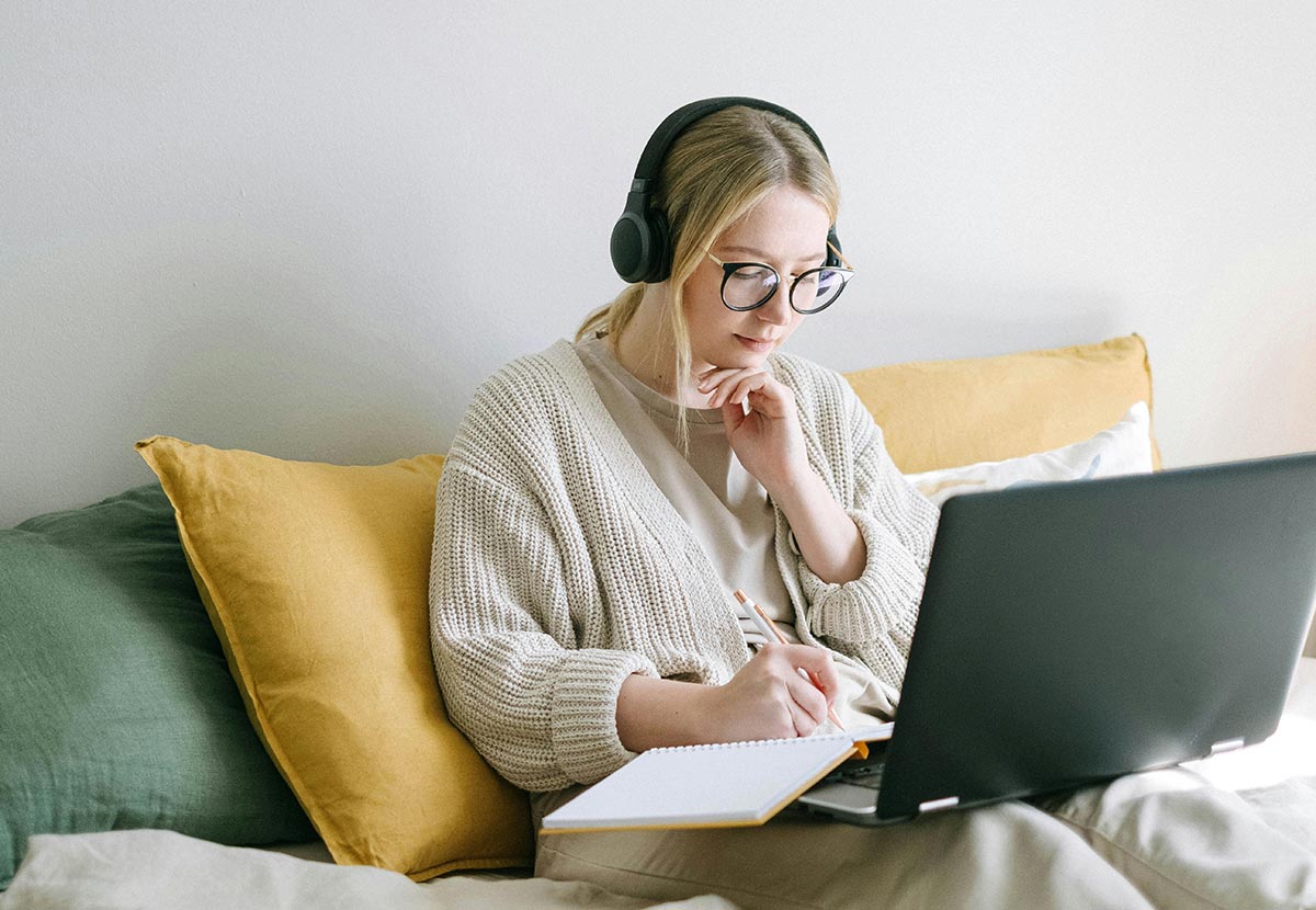 A person wearing headphones, sitting on a bed with pillows, taking notes while working on a laptop.