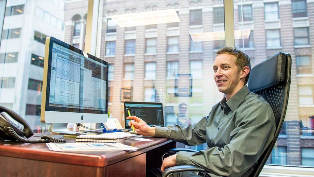 A person sitting at a desk in an office, working on a computer with large windows showing a cityscape in the background.