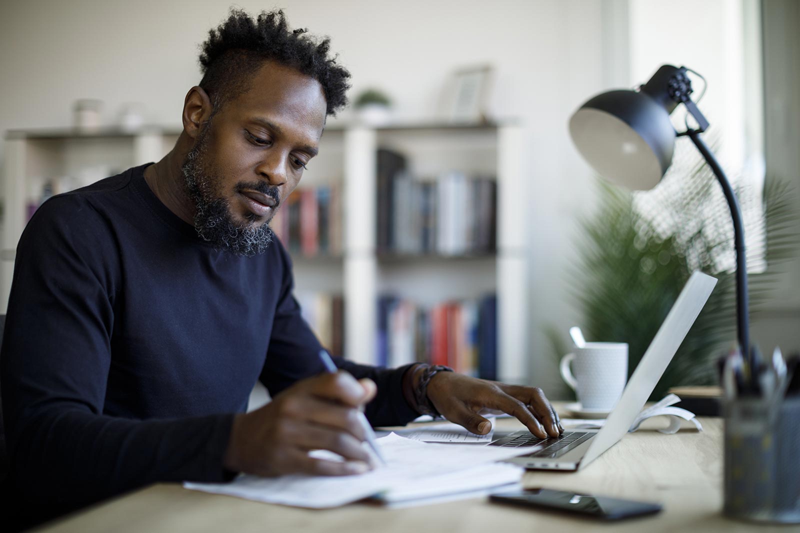 A person sitting at a desk, writing notes while using a laptop, with bookshelves in the background.