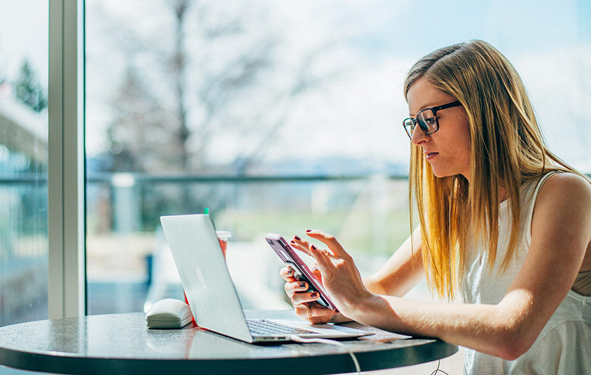 A young woman with blonde hair and glasses sits at a table, focused on her phone, with a laptop and drink beside her. A large window behind her shows an outdoor view.