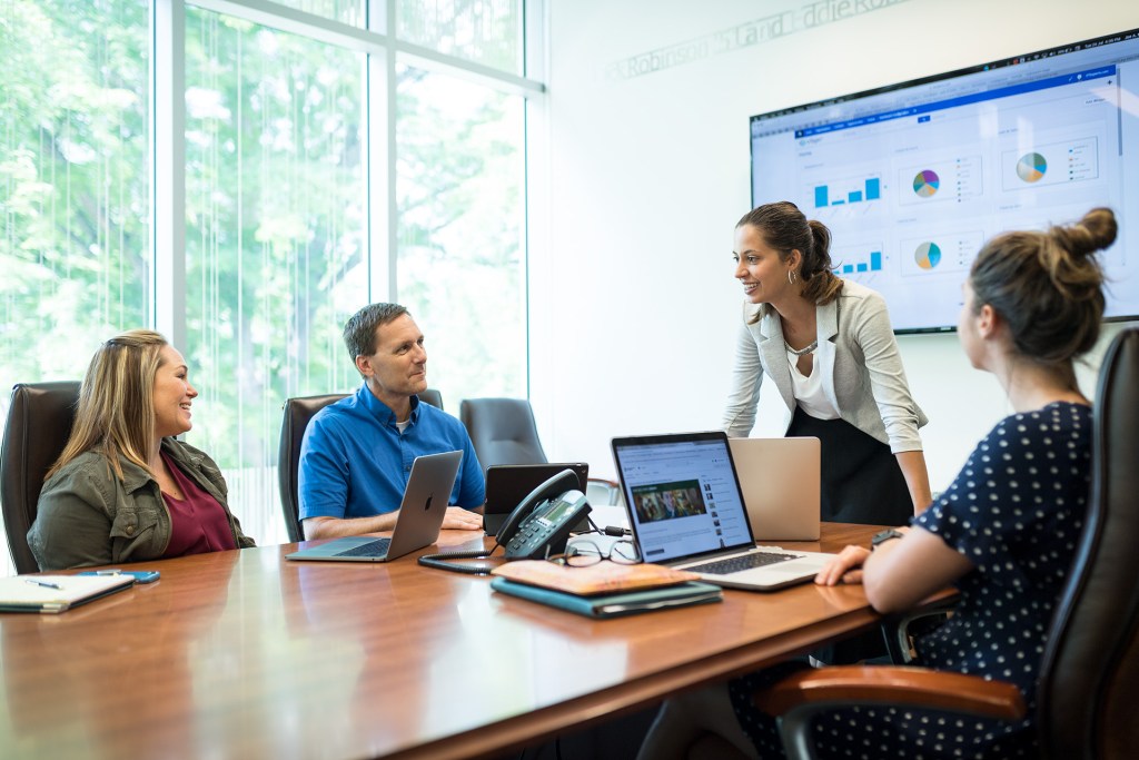 A group of people in a meeting room, engaged in discussion around a table with laptops, as one person stands and presents data displayed on a screen.