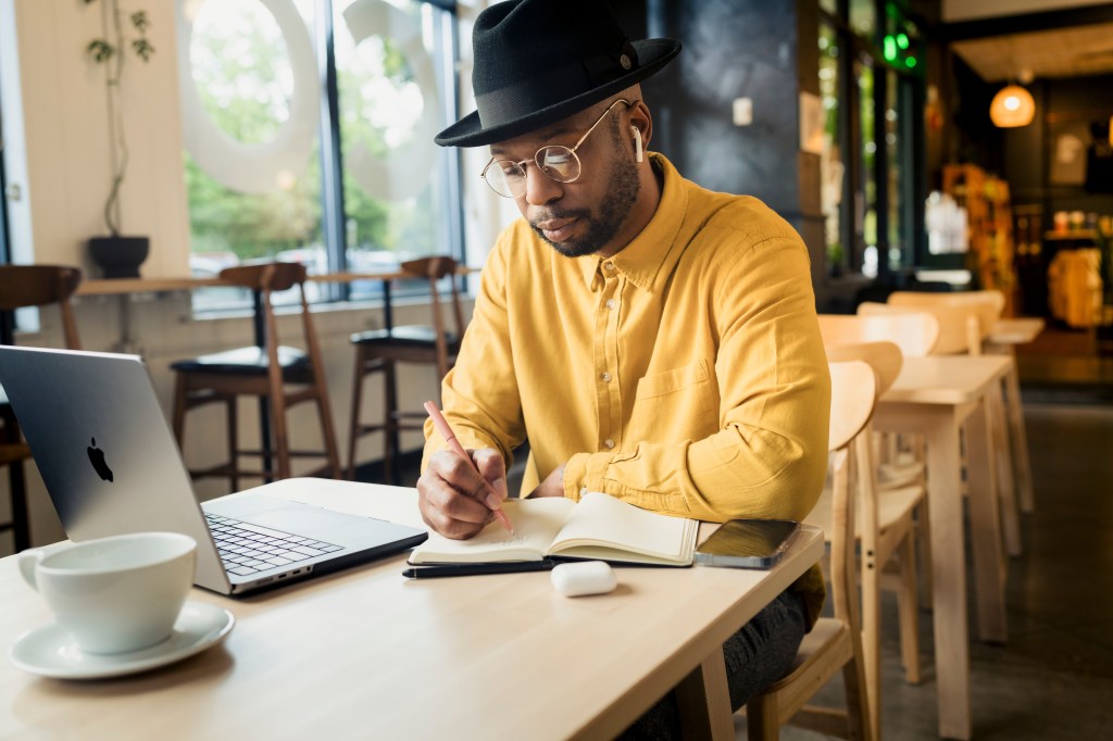 A man at a table in a coffee shop studying, writing in a notebook.