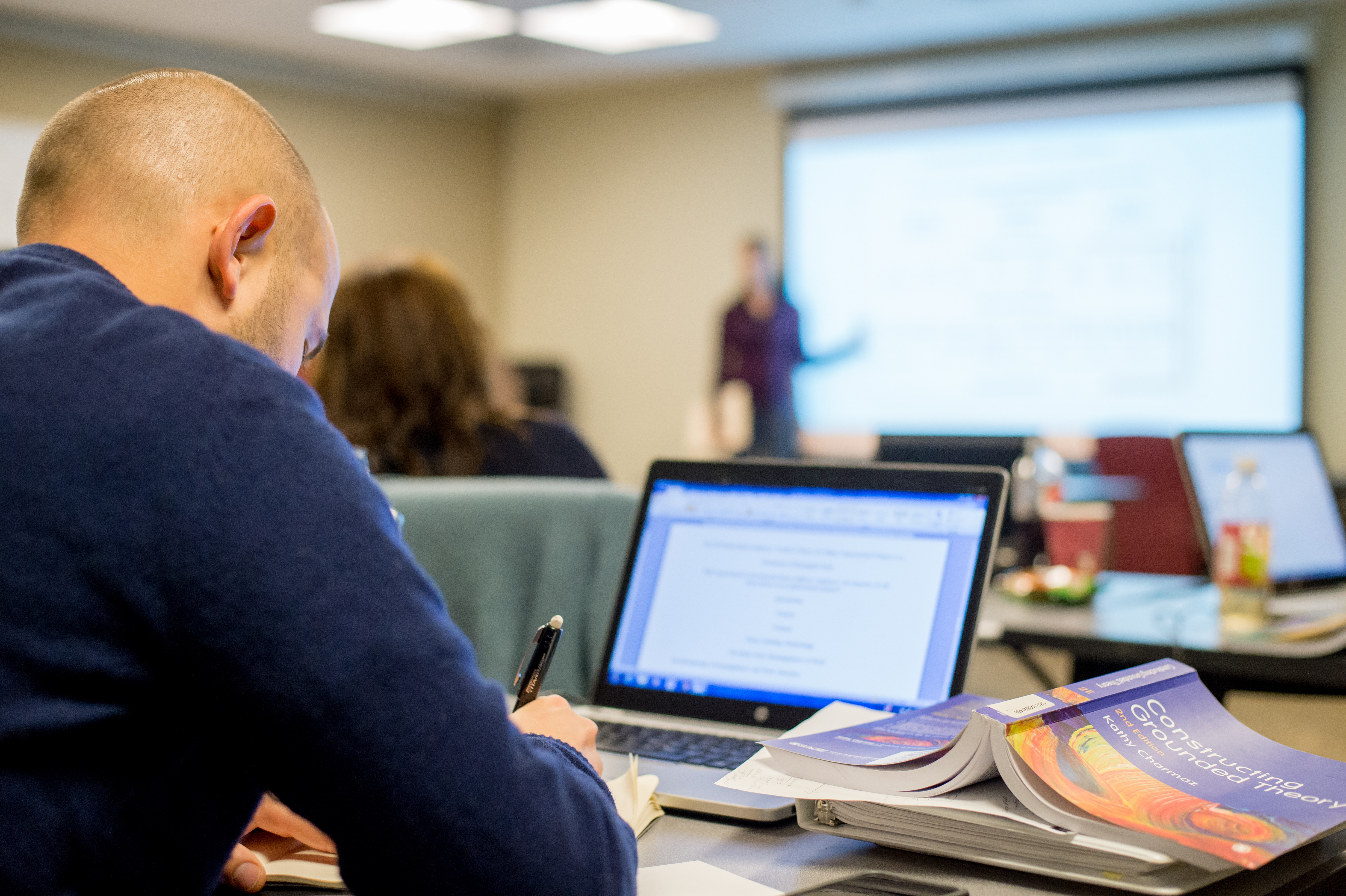 A person working on a laptop with a textbook nearby, taking notes with a pen.