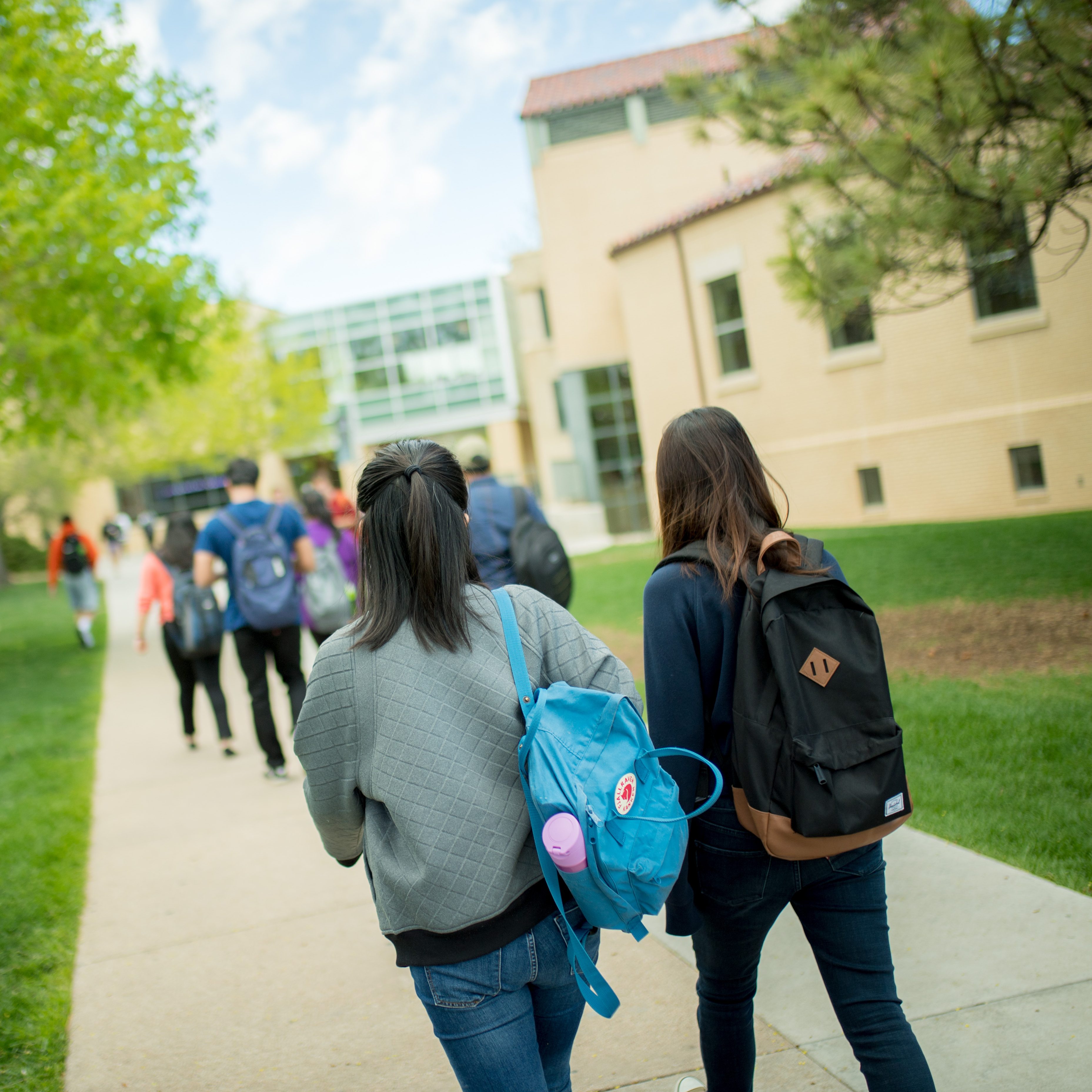 Two students with backpacks on walk down a sidewalk on campus.