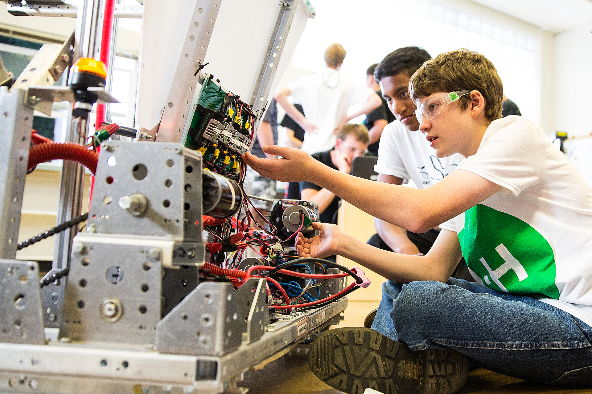 Students working on a complex robotic machine, focusing on wiring and components, with others observing in the background.