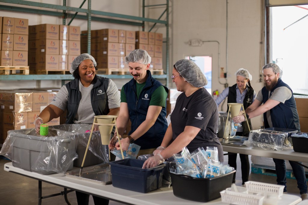 A group of individuals wearing hairnets and uniforms work together in a warehouse, assembling and packaging items. They are smiling and engaged in teamwork, with shelves of boxes and equipment visible in the background.