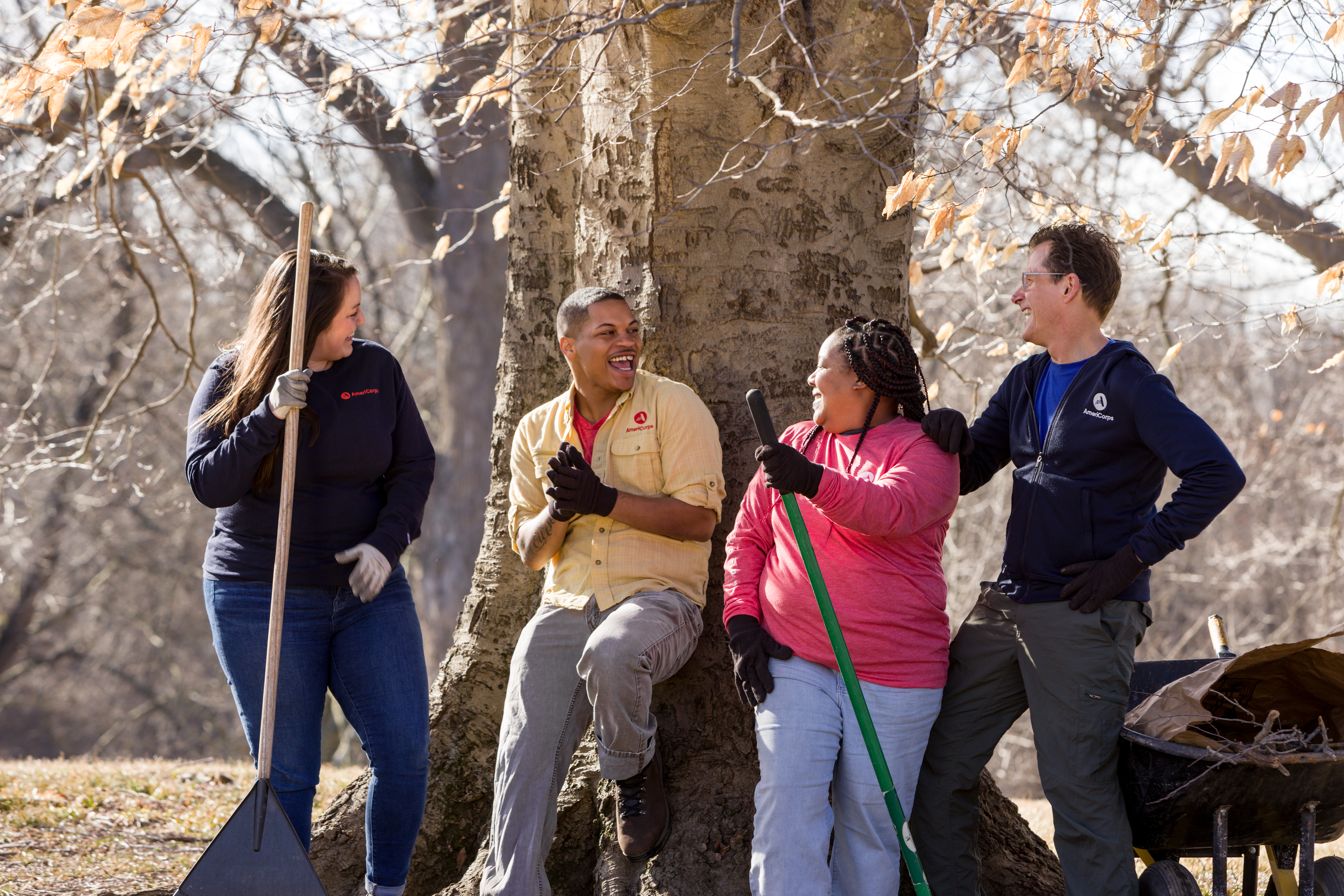 A group of four people stand and lean against a tree, smiling and chatting, while holding rakes and tools, suggesting they are engaged in an outdoor activity or community work.