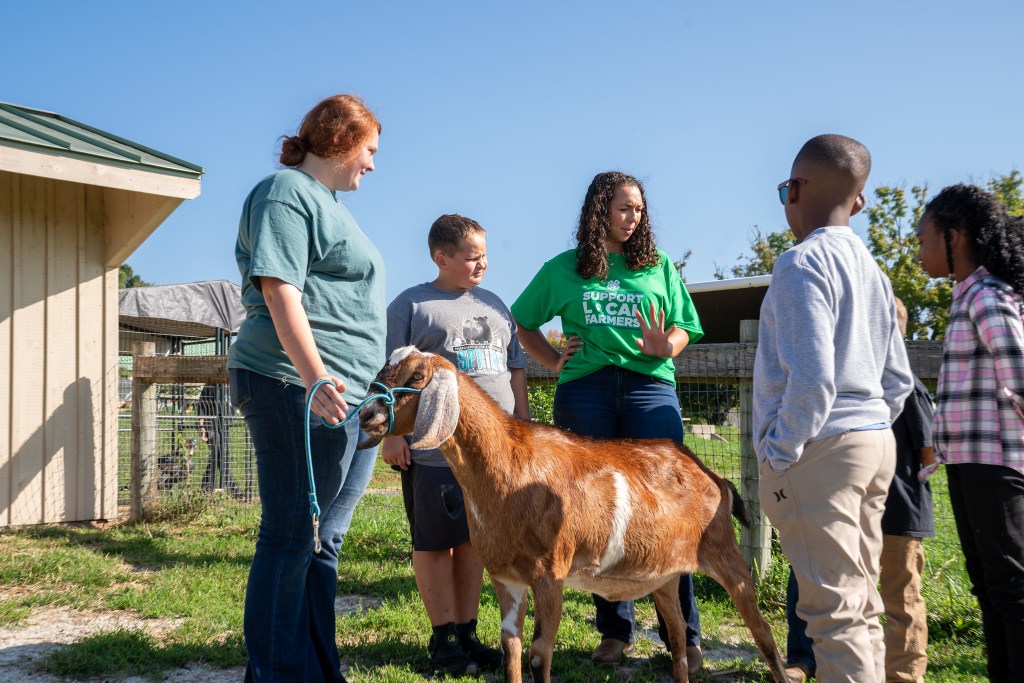 A group of people, including children, standing outside with a goat as one person holds the goat's leash while engaging in conversation.