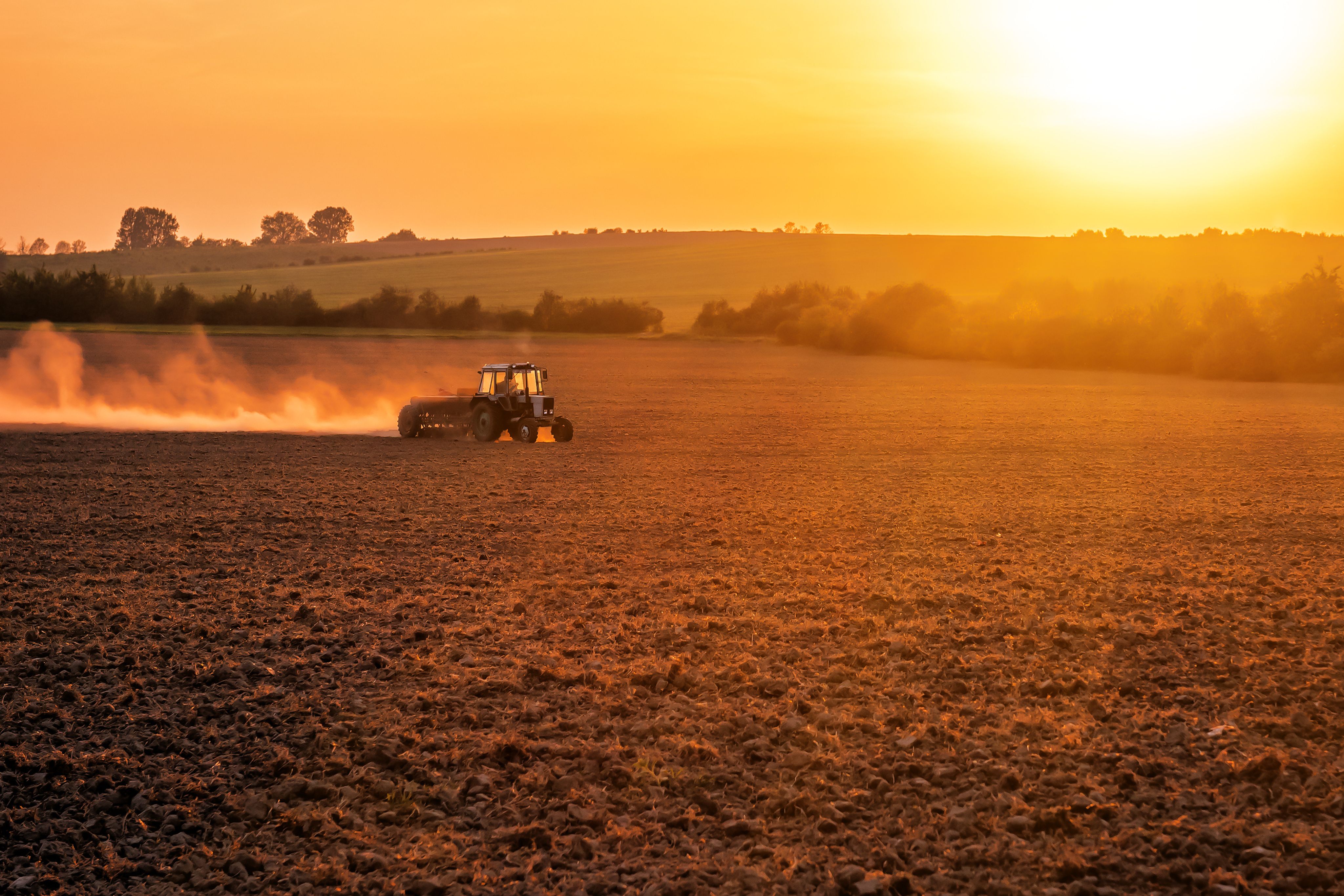 A tractor plowing a vast field with dust rising behind it during a beautiful sunset, creating a silhouette against the golden sky.