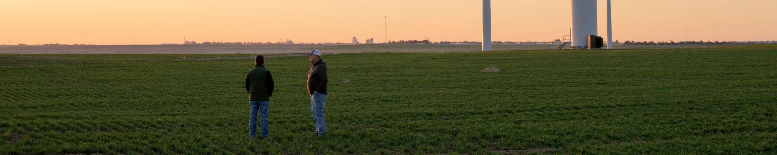 Two men stand in a vast green field at sunset, discussing or surveying the land with wind turbines visible in the background.