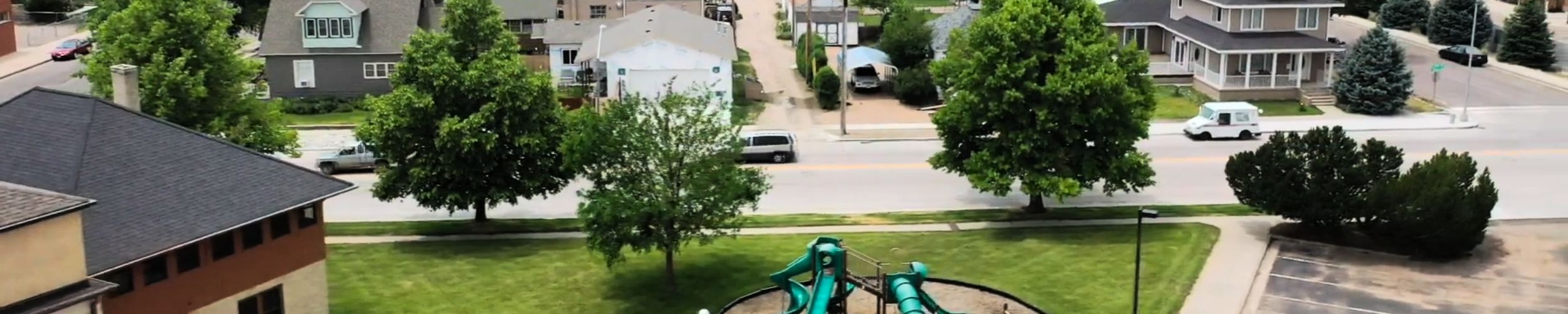 Aerial view of a neighborhood playground with green slides and surrounding homes, showcasing a residential area with mature trees, grass, and parked vehicles, conveying a sense of community and local environment.