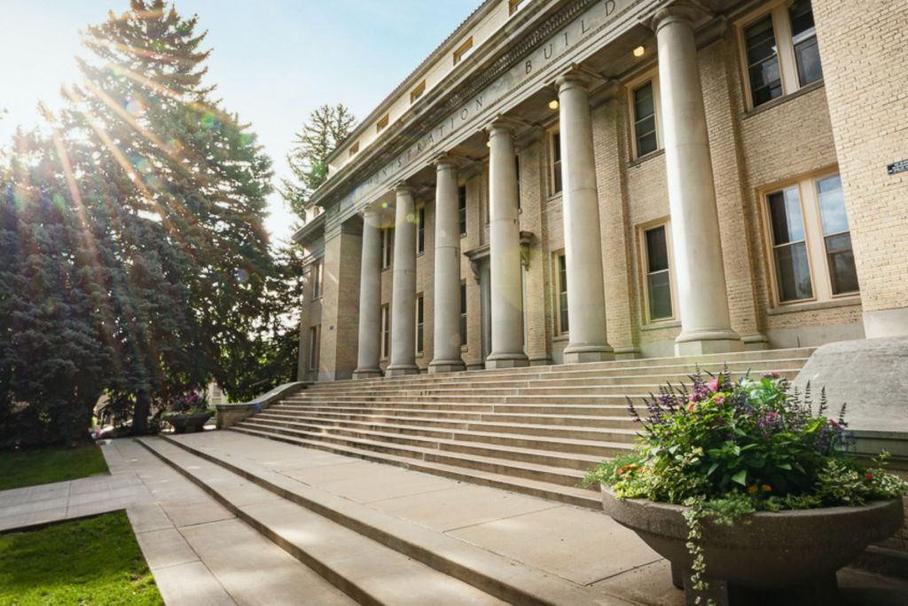 The historic Administration Building at Colorado State University with sunlight streaming through trees, highlighting its classic columns and steps.