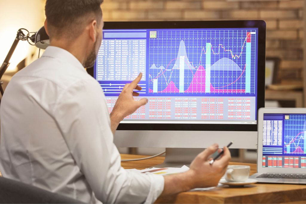 A man in a white shirt is analyzing graphs and data charts on a large desktop screen, discussing financial trends in an office setting.