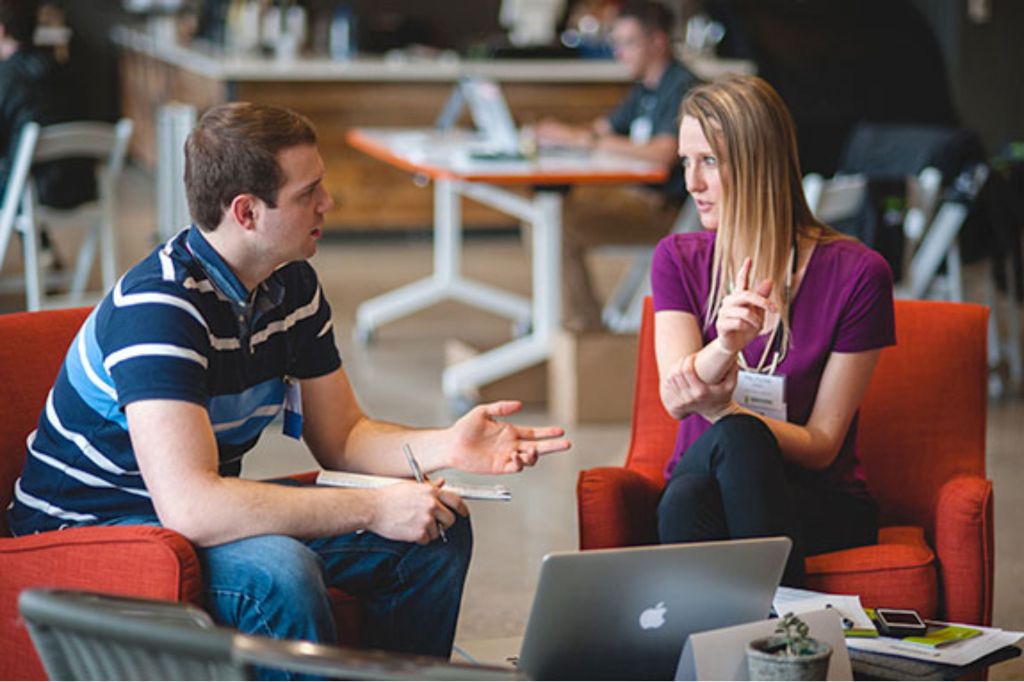 Two people in a casual setting discussing something while seated on orange chairs, with laptops and notebooks in front of them.