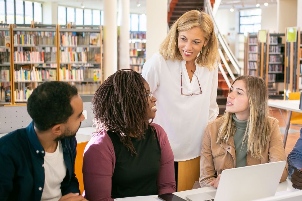 A diverse group of students and a teacher having a discussion in a library, with shelves of books visible in the background.